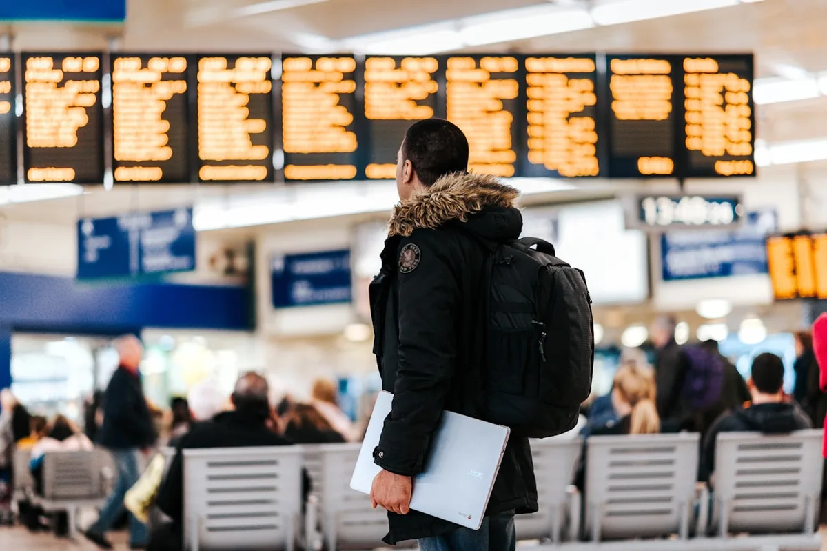 공항 터미널에서 배낭을 멘 여행자가 출발 안내 전광판을 바라보는 모습으로, 여행지에 따라 유리한 트래블카드가 다르다는 점을 표현한다